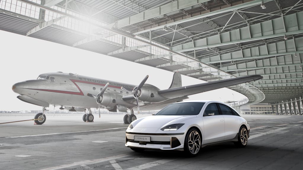 Sleek white electric car parked near a vintage aircraft in an open hangar.